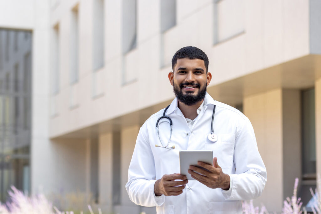 Portrait of a young male doctor, nurse, standing outside the hospital, holding a tablet and looking into the camera